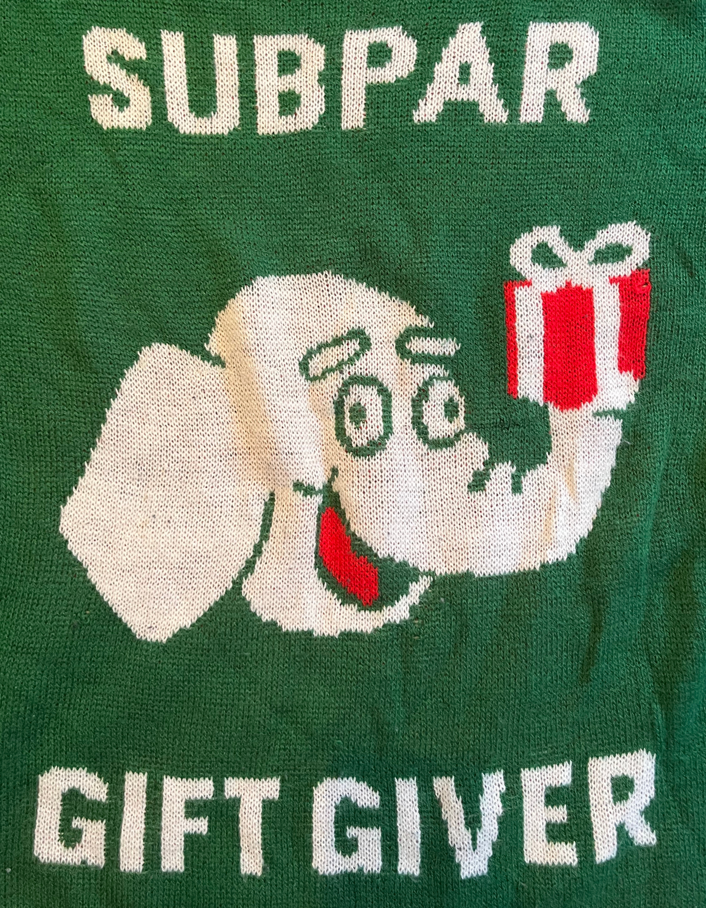 Close up image of green and red sweater featuring elephant holding a gift with text reading subpar gift giver on white background.