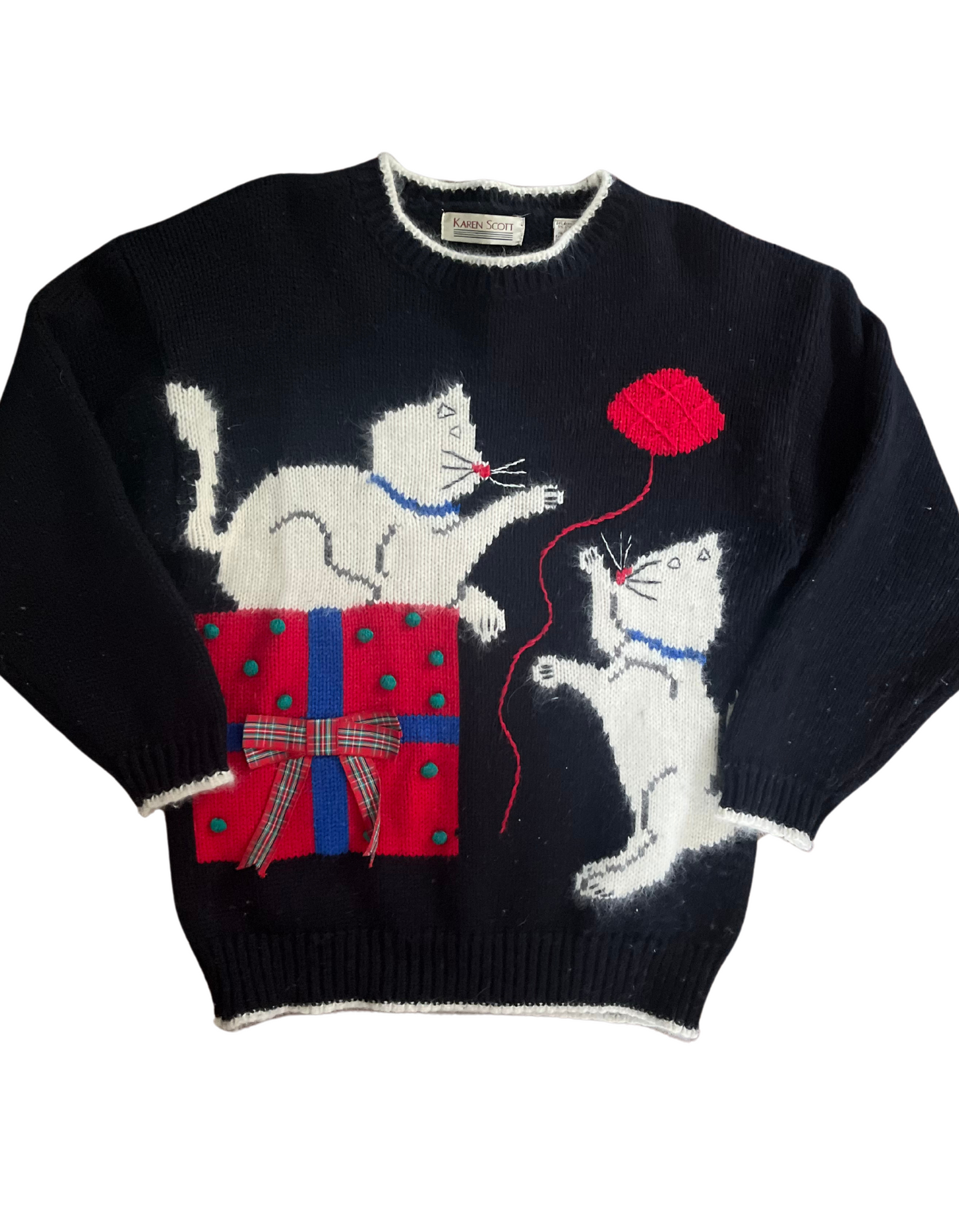 Front image of vintage holiday sweater featuring 2 white kitty cats playing with yarn next to a christmas gift on light background.